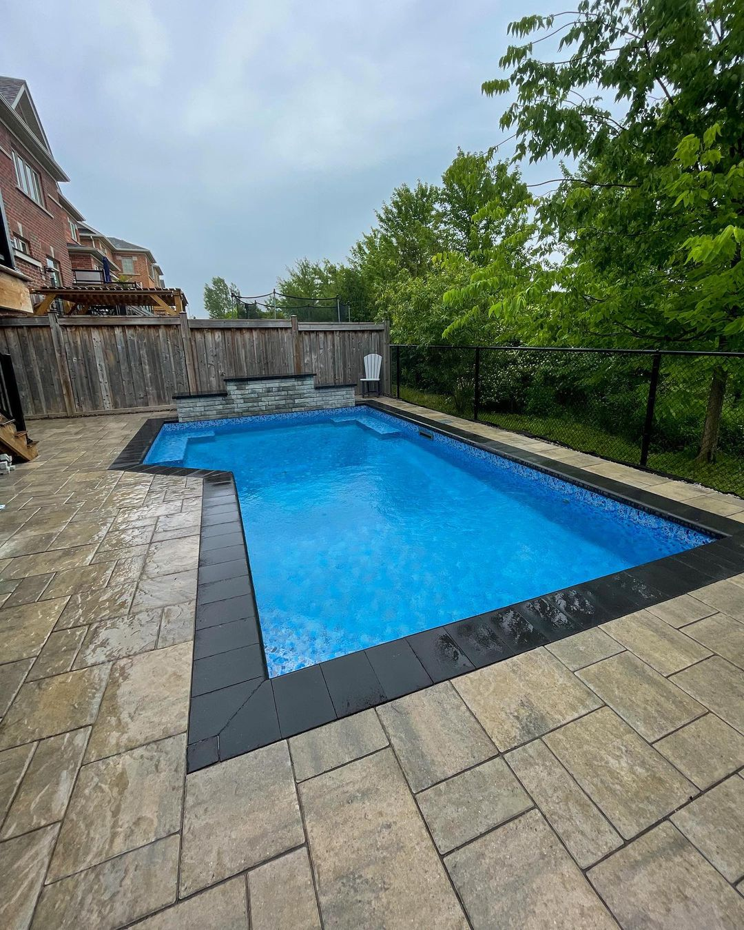 A backyard swimming pool with blue water, surrounded by a stone-tiled deck and black border tiles. There's a wooden fence, some green trees, and a cloudy sky.