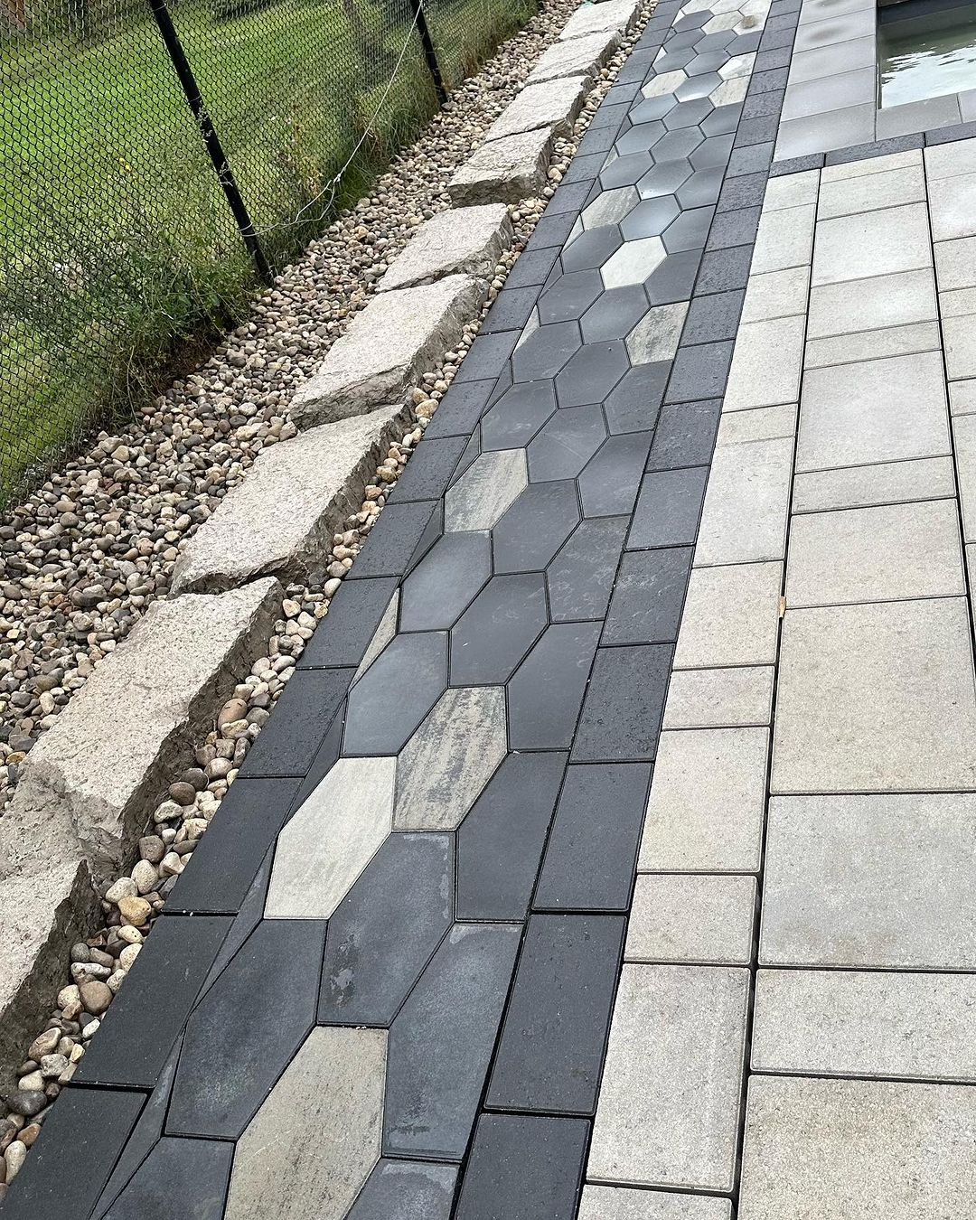 Close-up of a paved pathway with different types and colors of stones and bricks, bordered by white gravel and rocks, next to a green grassy area and black fence.