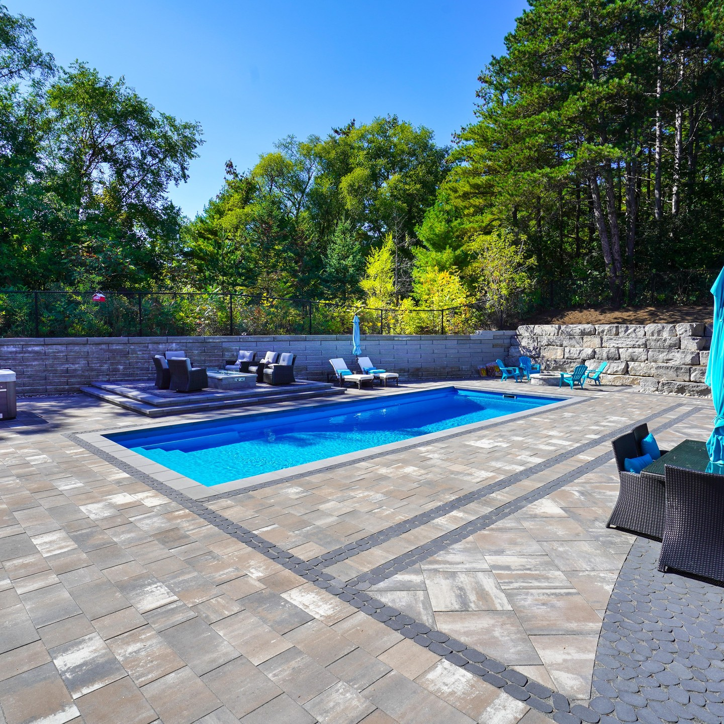 Backyard with a swimming pool surrounded by a stone deck, outdoor furniture, and a backdrop of green trees and a clear blue sky.