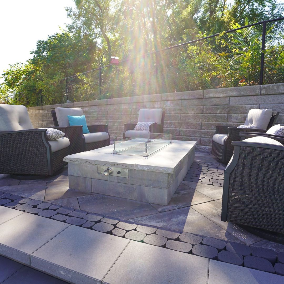 Outdoor patio with cushioned chairs around a fire pit table, sunny day with sunlight rays and green trees in the background.