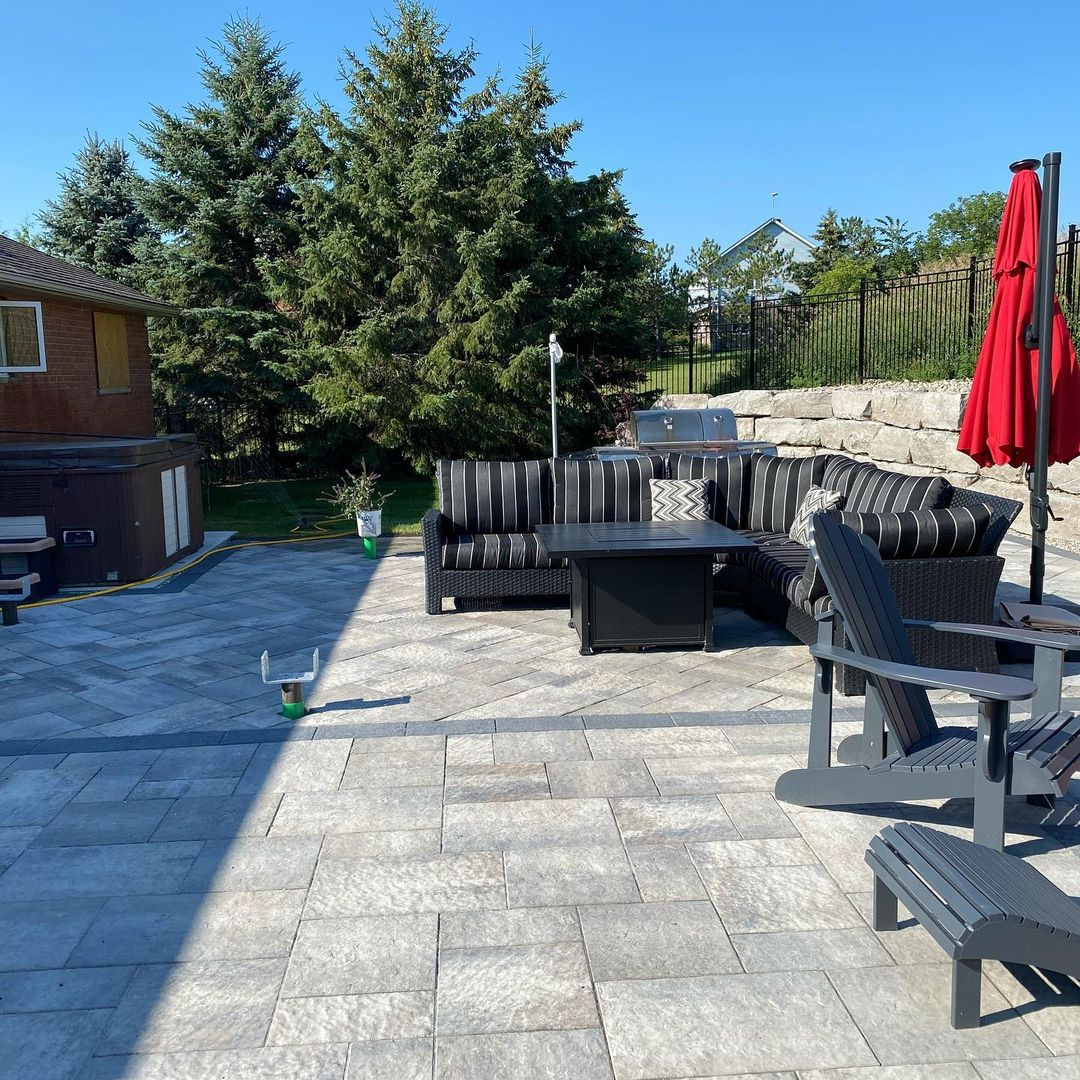 A backyard patio with light gray stone tiles, a black outdoor sectional sofa with black and white striped cushions, a black fire pit table, a gray Adirondack chair, a red patio umbrella, green trees, a brick building, a hot tub, and a stone retaining