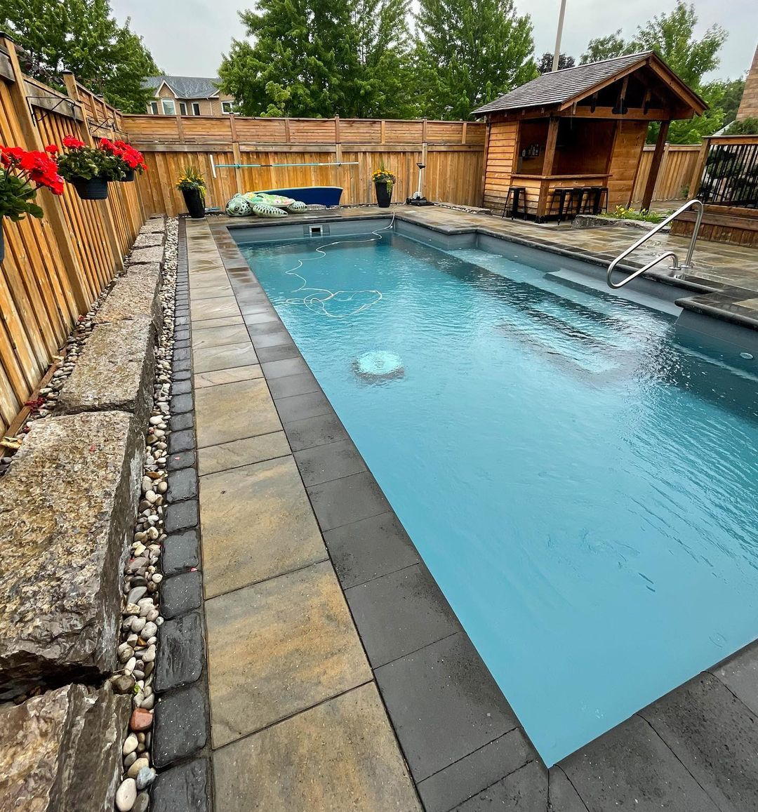 A backyard swimming pool with a hot tub, surrounded by stone and tile paving, a wooden fence, potted plants, and outdoor furniture including a small wooden shelter with stools.