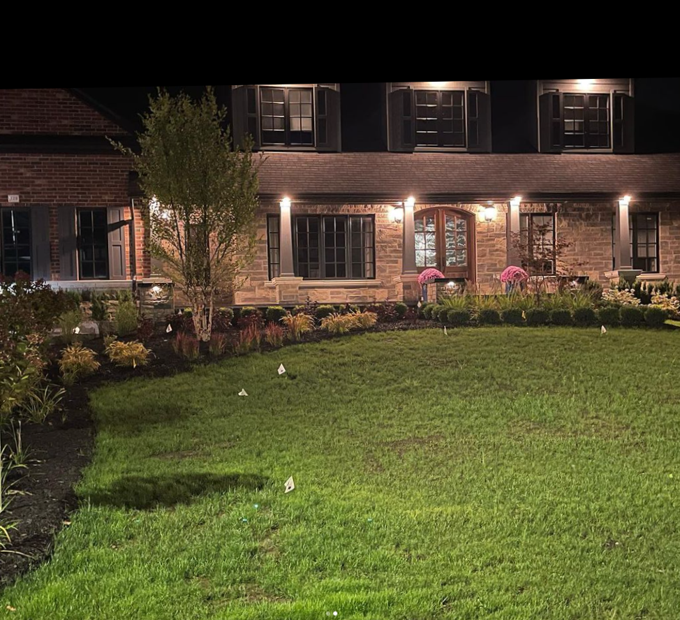 Nighttime view of a house with exterior lighting, brick and stone facade, multiple windows with shutters, and a well-maintained garden with grass, flowers, and small flags.