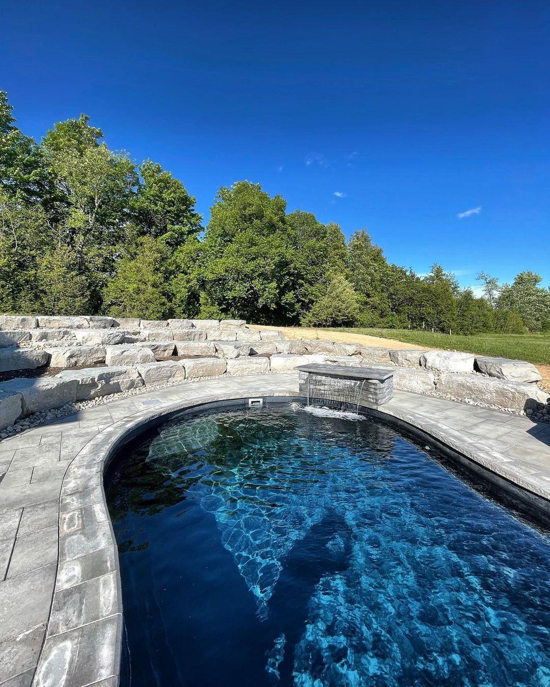 An outdoor swimming pool with stone edging and a waterfall feature, surrounded by green trees and a clear blue sky.