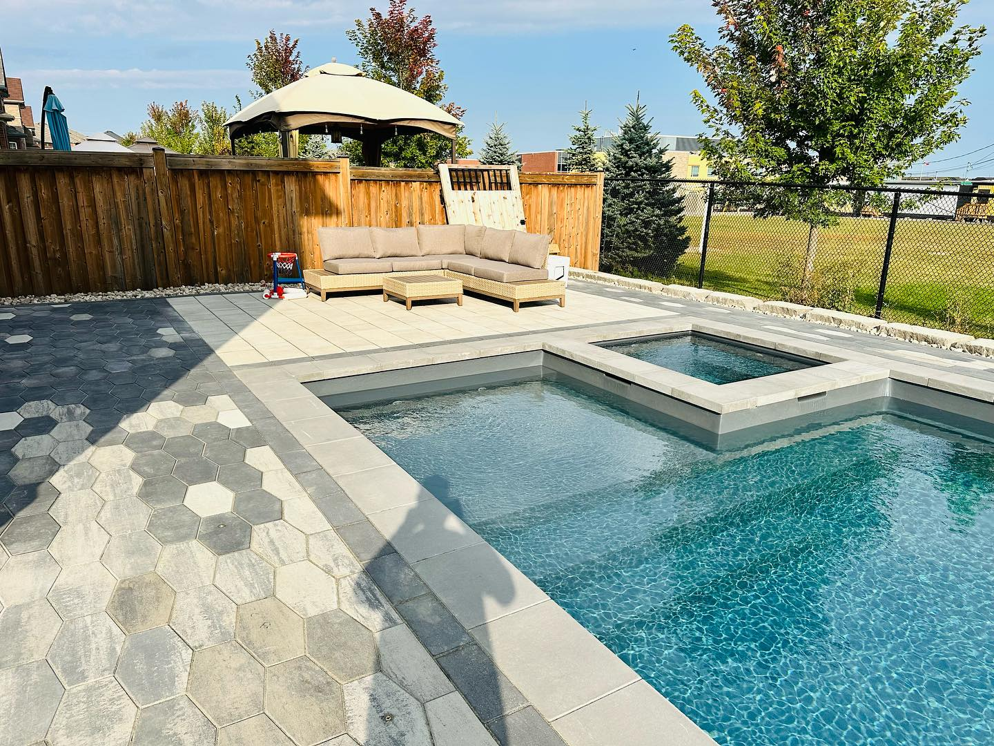 A backyard patio with a swimming pool and hot tub, decorated with outdoor furniture including a sectional sofa with cushions, a small table, and an umbrella-covered seating area. There is a wooden fence and some trees in the background.