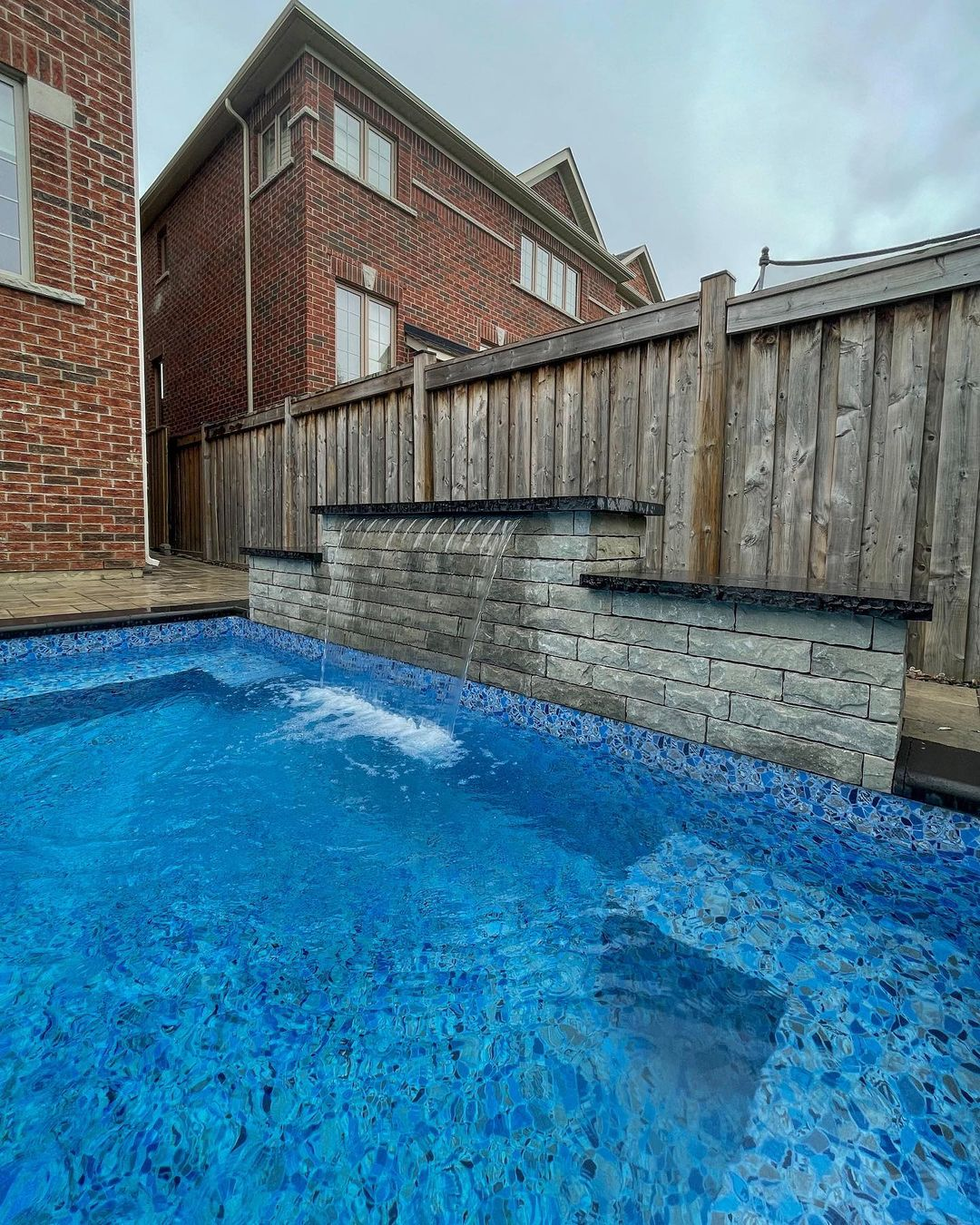 Backyard swimming pool with a waterfall feature, surrounded by a wooden privacy fence and adjacent brick house.