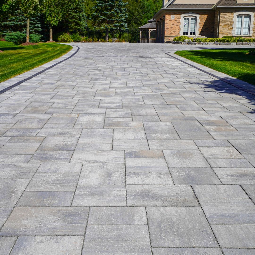 A paved driveway with gray stone tiles leading to a house, surrounded by green grass and trees.