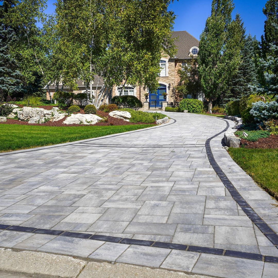 A paved driveway leading to a house with a brick exterior, surrounded by well-maintained landscaping, trees, and shrubs.