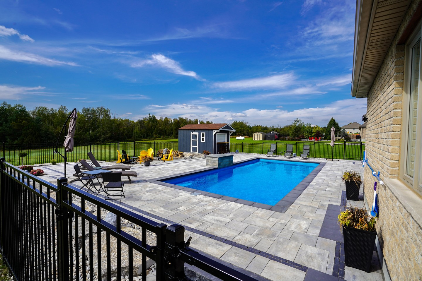 Residential backyard with a swimming pool, poolside chairs, umbrellas, a small blue shed, and a large grassy area with trees under a blue sky with clouds.
