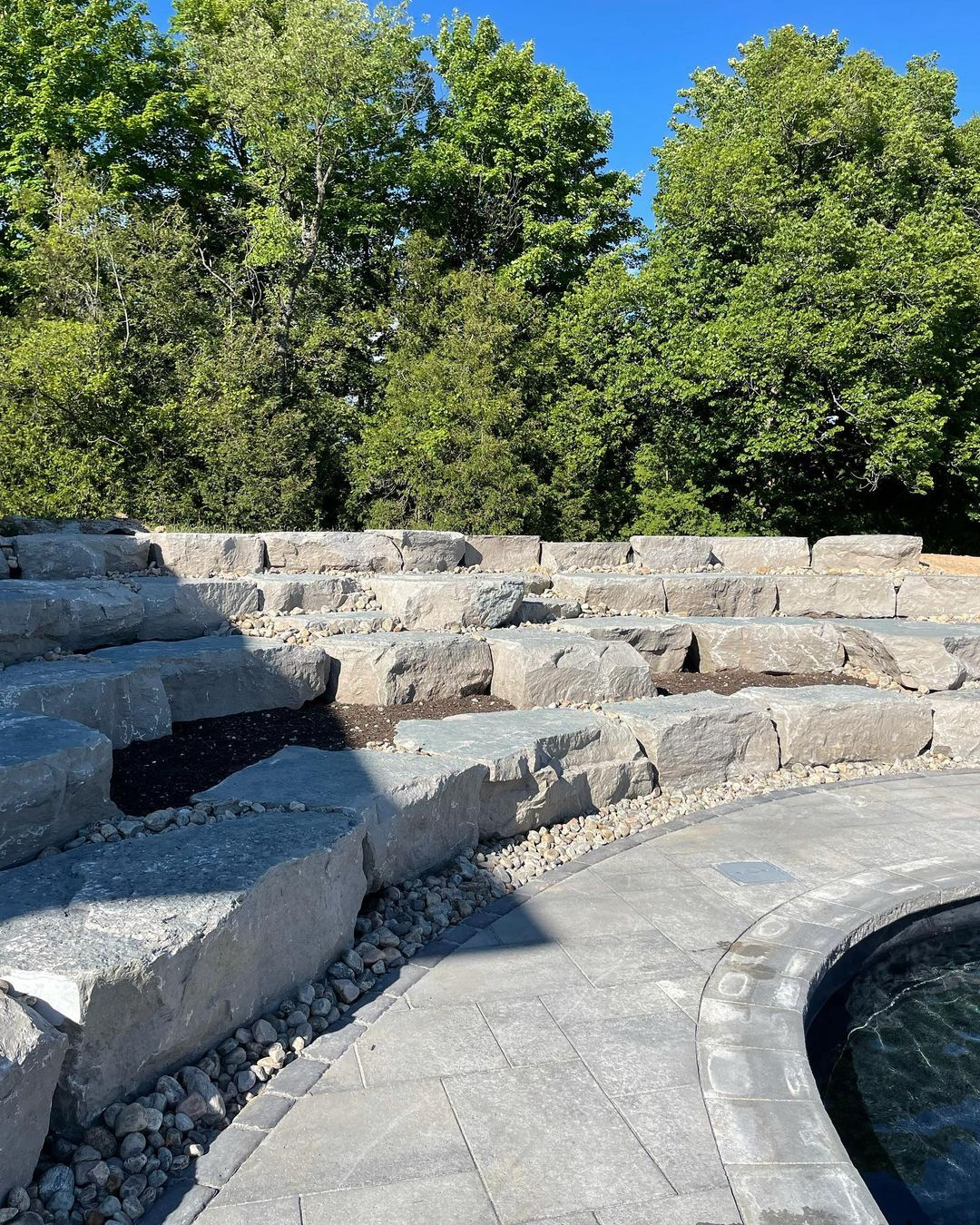 Stone amphitheater-like steps surrounded by trees and a curved paved area with a water feature, under a clear blue sky.