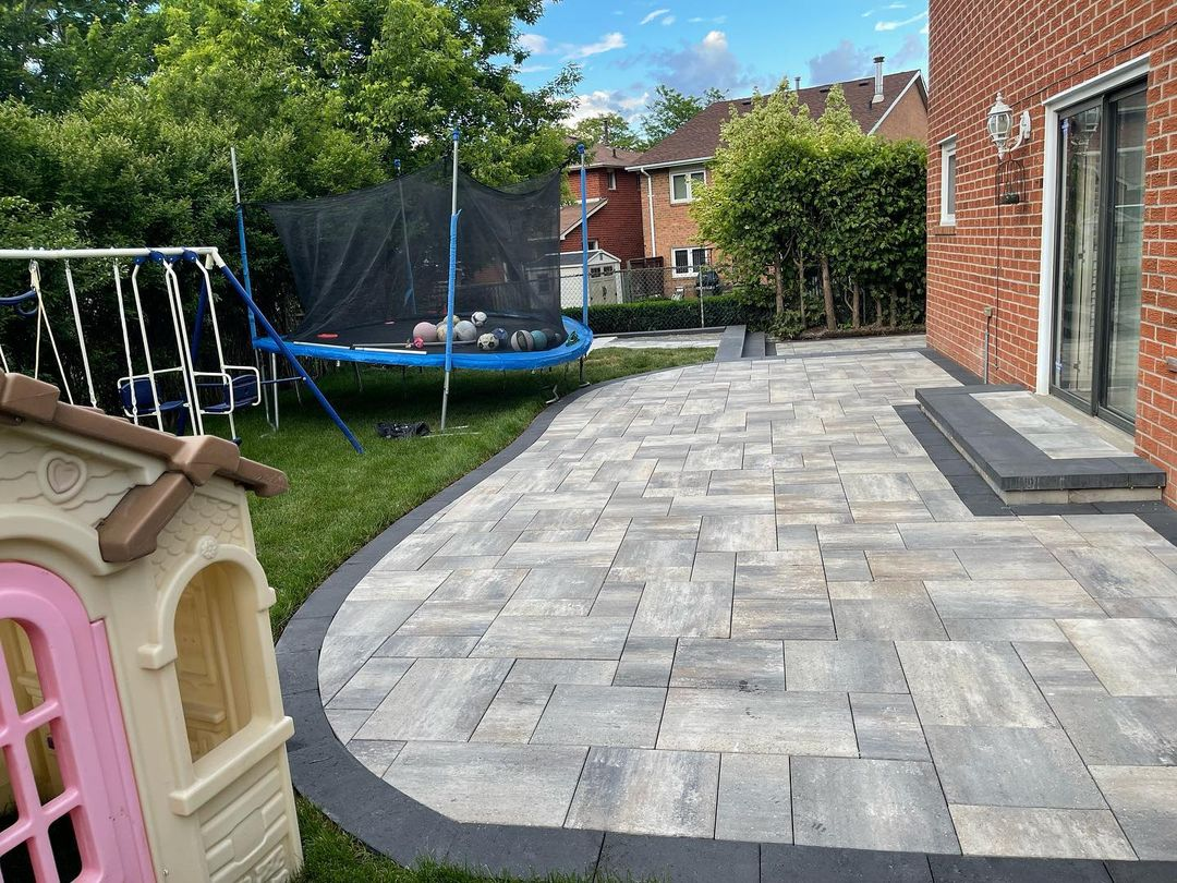 Backyard patio with a brick house, a black sliding door, and a small set of steps. The patio is paved with grayish tiles, curving along a grassy area. There is a children's plastic playhouse on the grass, a swing set, and a trampoline with a safety n