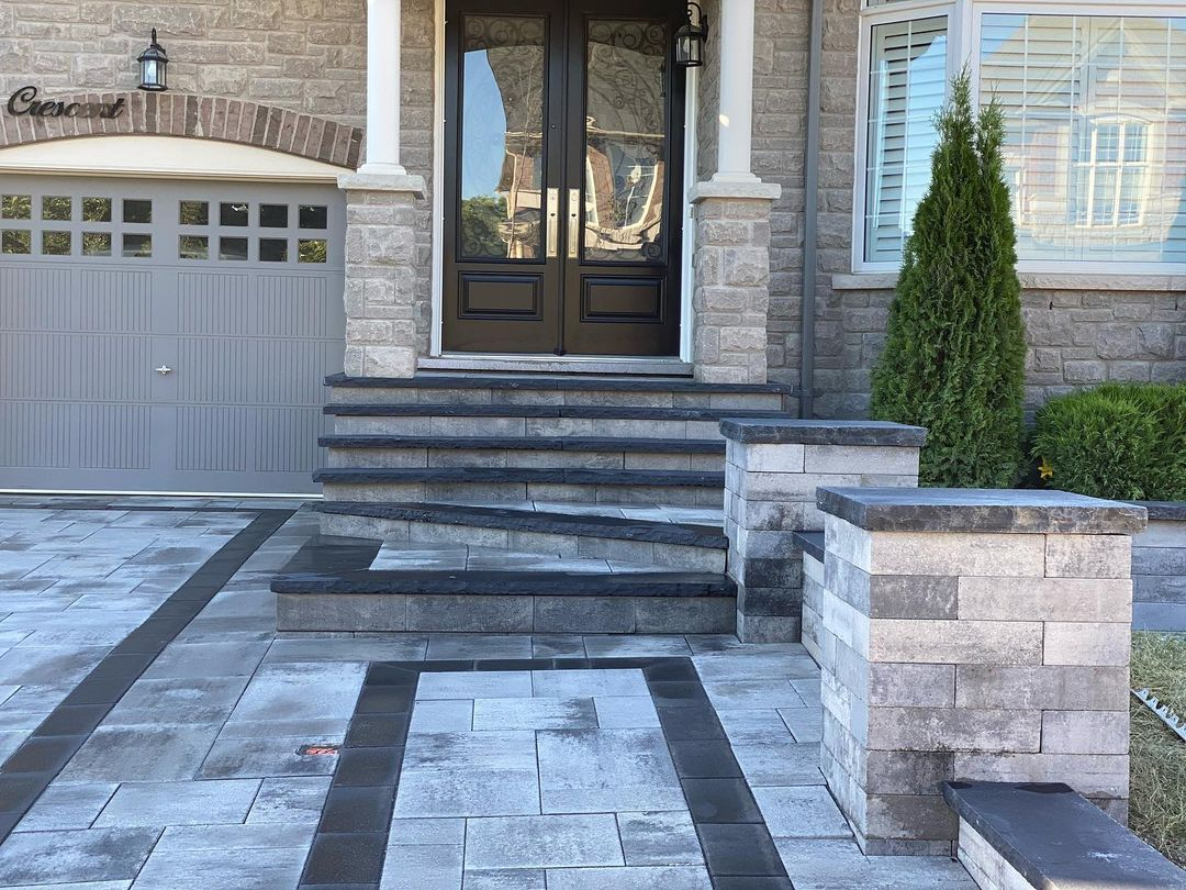 Concrete steps leading up to a black front door with glass window panels, flanked by stone pillars, with a garage door on the left and a large window with shutters on the right, landscaped with small shrubs and a tall evergreen tree.