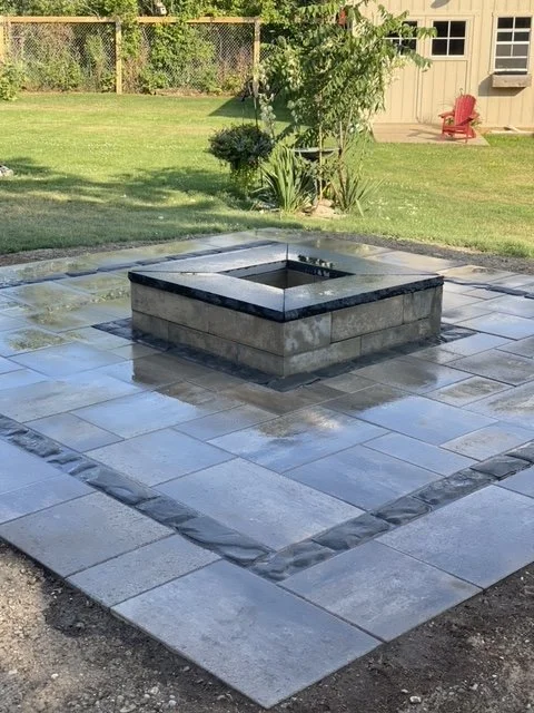 Small fire pit with a tiled patio and grassy backyard with plants and a shed.