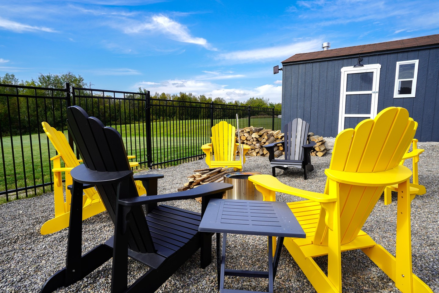 Outdoor seating area with black and yellow Adirondack chairs around a fire pit, gravel ground, a metal fence, a gray shed with a white door, and wood logs