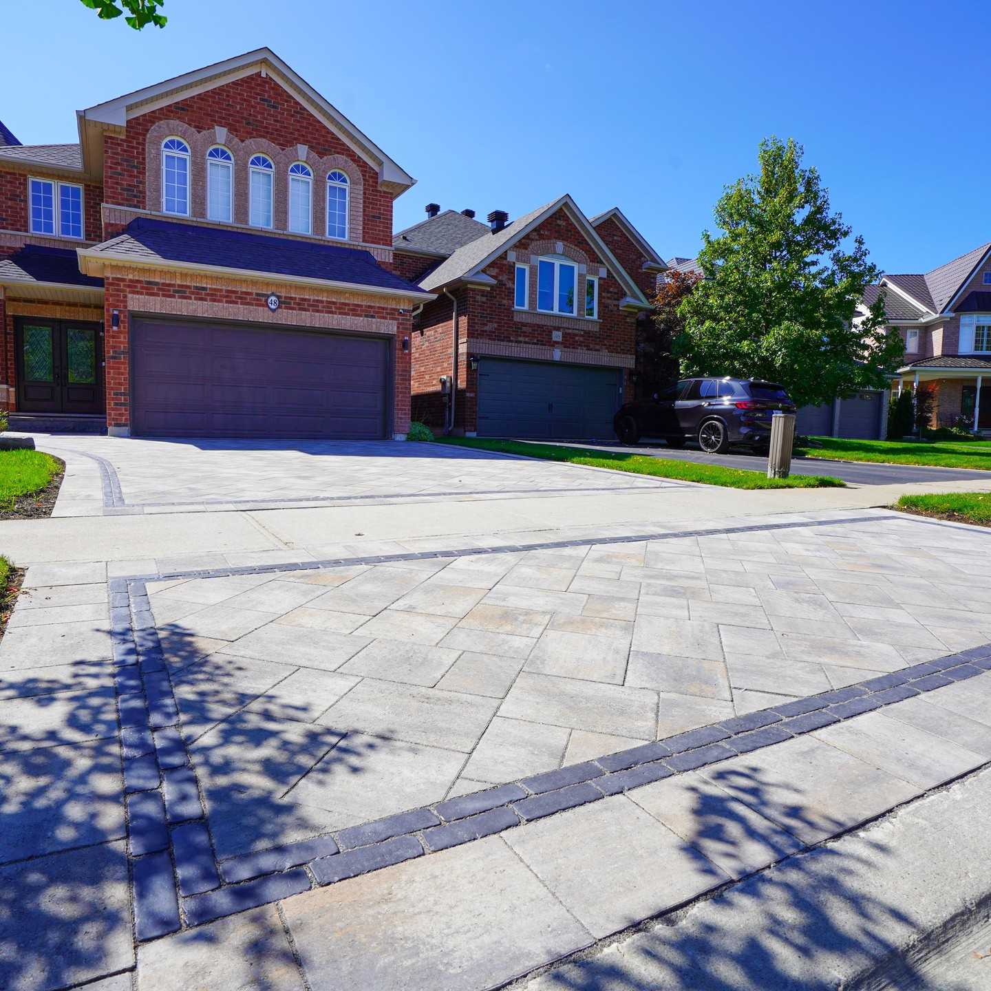 A suburban house with a brick exterior, two-car garage, driveway, trees, and a black car parked on the street.