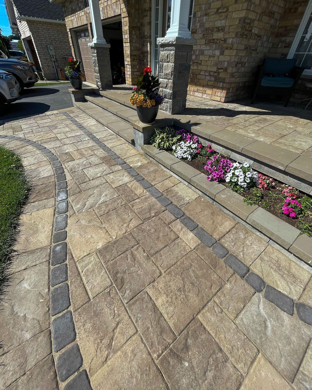 Residential front porch with stone steps, flower pots, and flower beds near a brick house. The porch has potted plants, and there are colorful flowers in the garden bed adjacent to the walkway. Cars parked in driveway on left side.