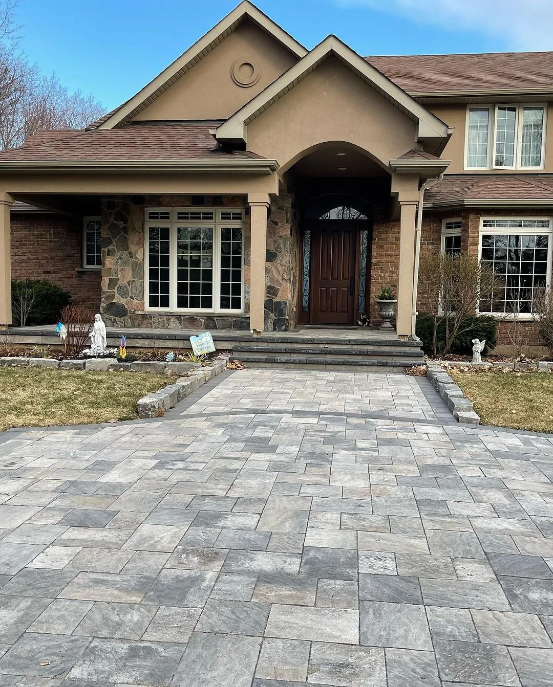 Front view of a large house with a stone and brick exterior, a dark wooden front door, stone steps leading to the entrance, a paved driveway and small landscaped garden with garden ornaments.