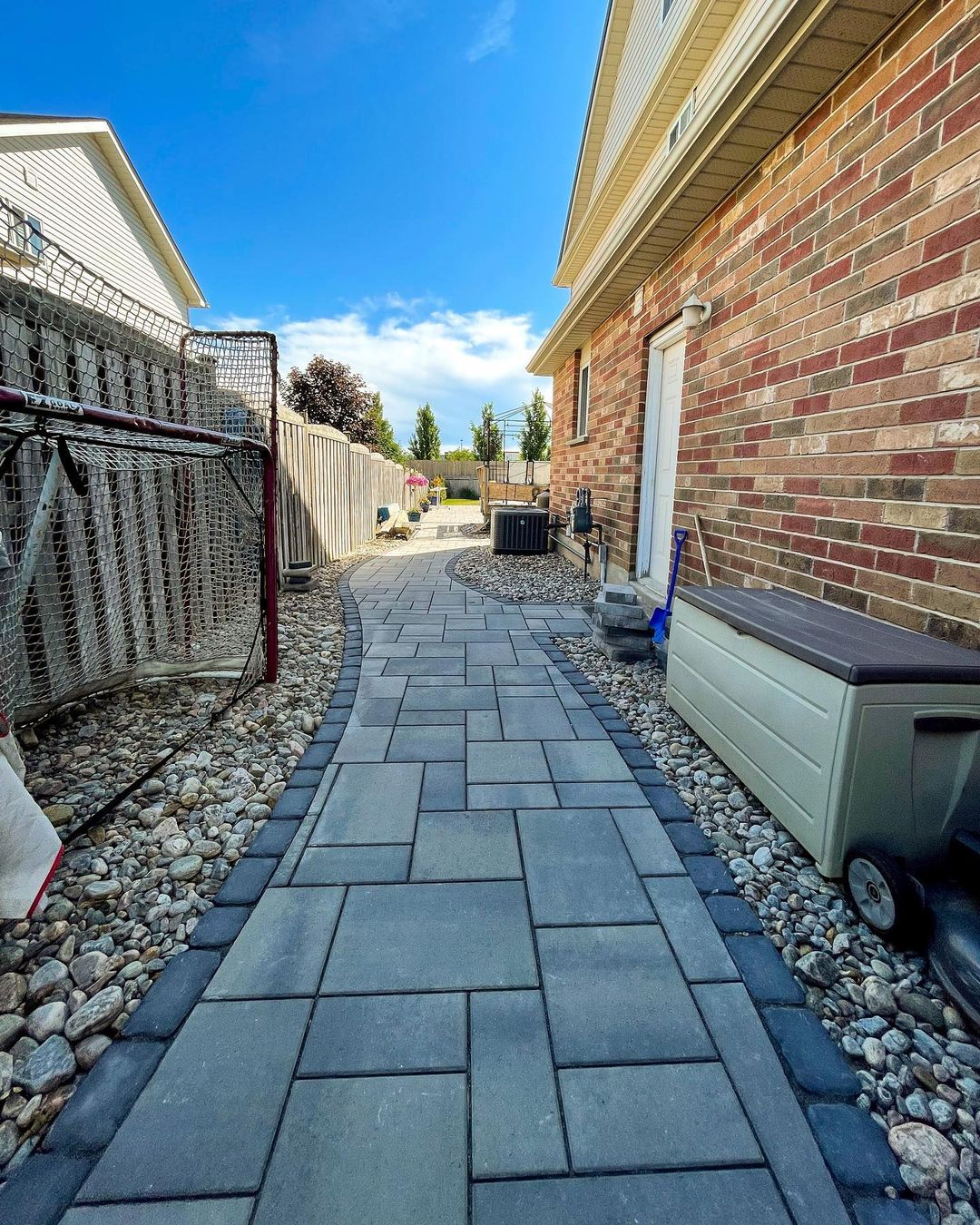 A backyard pathway with pavers, bordered by river rocks, runs alongside a brick house under a bright blue sky with some clouds. There are outdoor storage containers, a snow shovel, and a bench along the path.