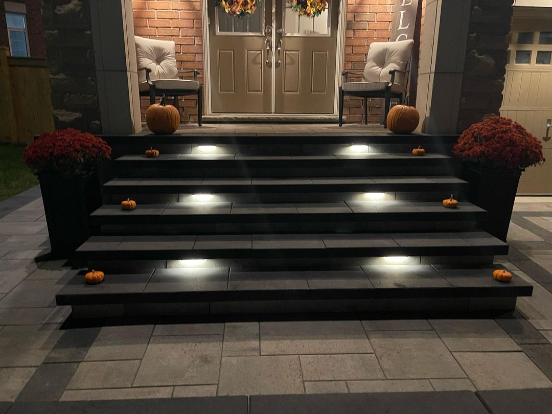 Front porch with black stairs decorated with small pumpkins, leading to a double front door. Two white chairs with cushions sit on either side of the door, with pumpkins placed beside both chairs. Red potted flowers are on the top of the stairs.