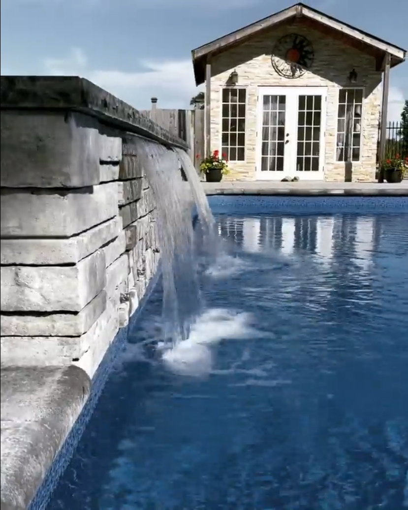 A backyard pool with a small waterfall feature made of gray stone, and a white shed or pool house with French doors and large windows in the background.