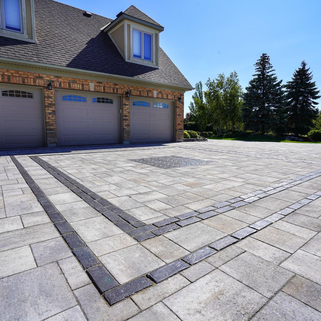 A brick and siding garage with three closed gray doors, and a concrete driveway with decorative dark border lines, surrounded by trees and green grass.