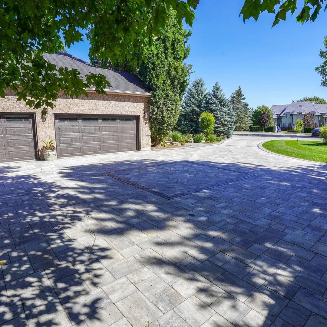 A paved driveway leading to a garage with a brick house, surrounded by trees and greenery on a sunny day.