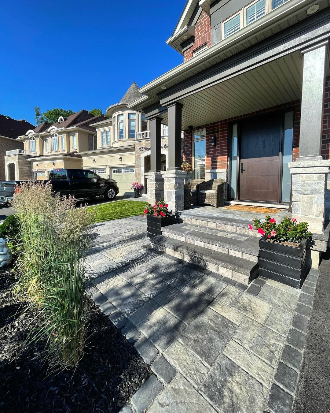 Front view of a well-maintained residential house with a brick and stone exterior, a porch with chairs, potted plants, and a paved walkway leading to the front door, under a bright blue sky.