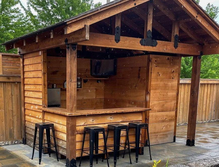 Wooden outdoor bar with three black stools, a TV, and a mini refrigerator, situated on a paved patio with a wooden fence and trees in the background.