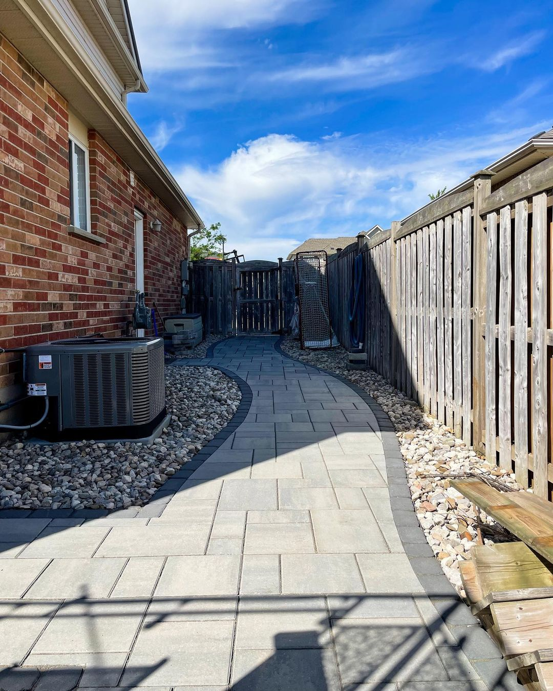 A winding brick patio path bordered by rocks, with a wooden fence on the right and a brick house on the left, under a partly cloudy blue sky.
