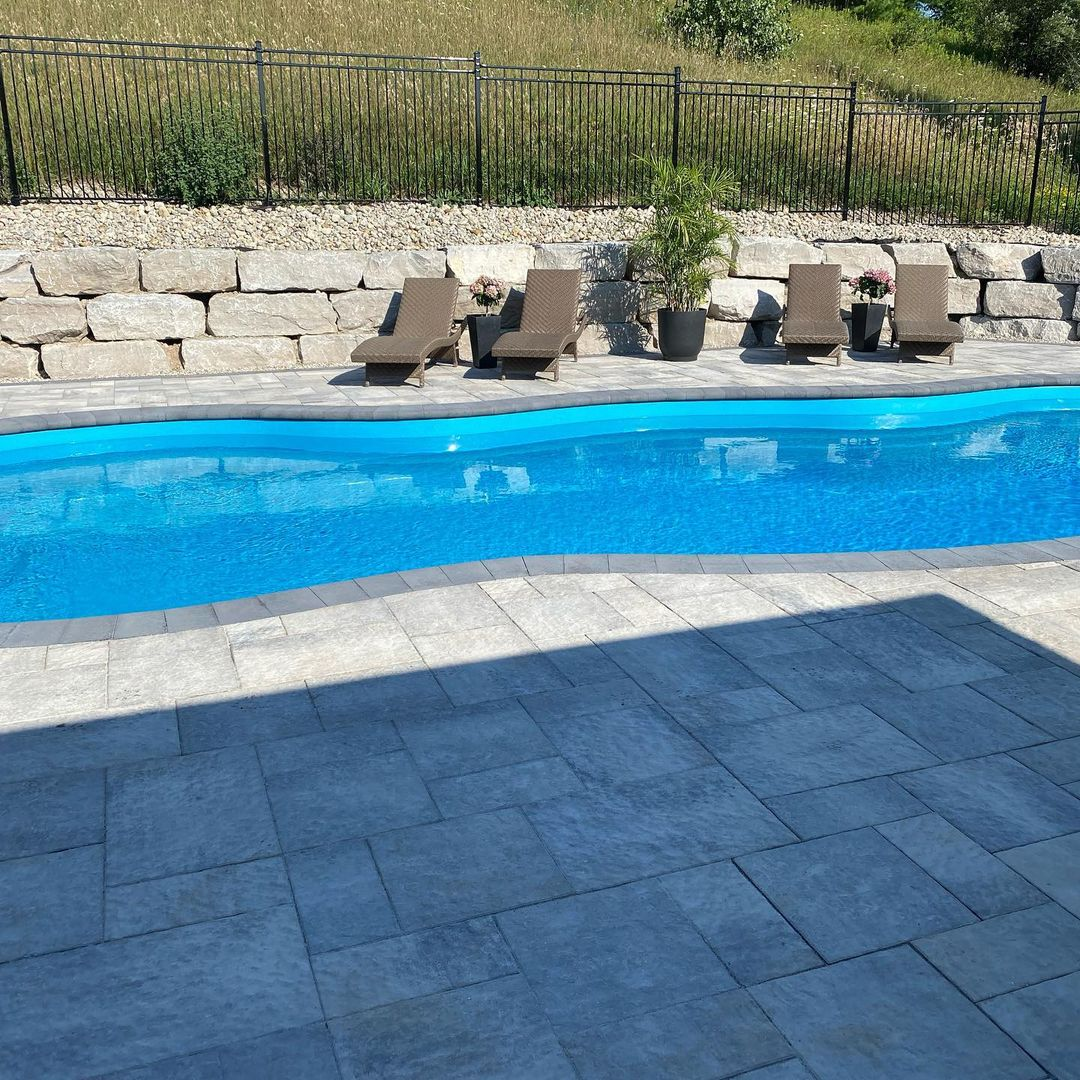 A backyard swimming pool with blue water, stone patio, four lounge chairs, potted plants, and a stone retaining wall with greenery behind it.