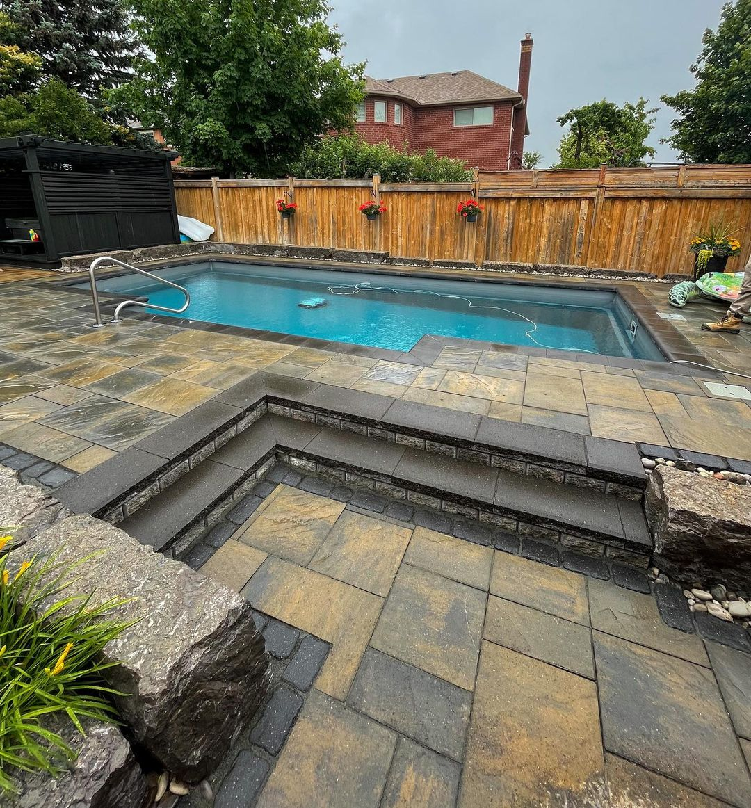 Backyard with a rectangular swimming pool, stone tiled patio, wooden privacy fence, potted flowers, and a house in the background.