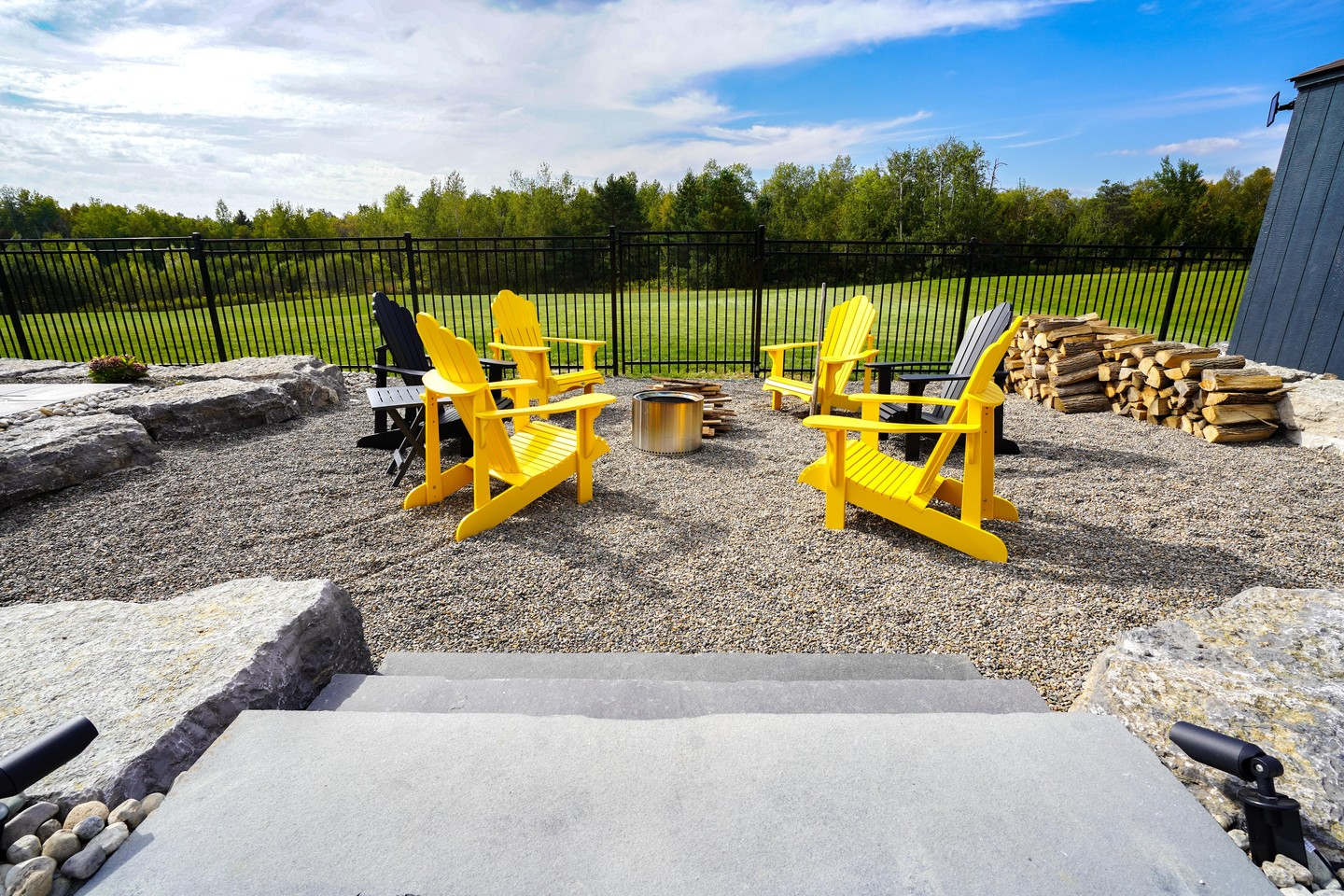 Outdoor patio with six Adirondack chairs in yellow and black, arranged around a fire pit, surrounded by gravel, stone, and a black metal fence, with stacks of firewood and a grassy field with trees in the background.