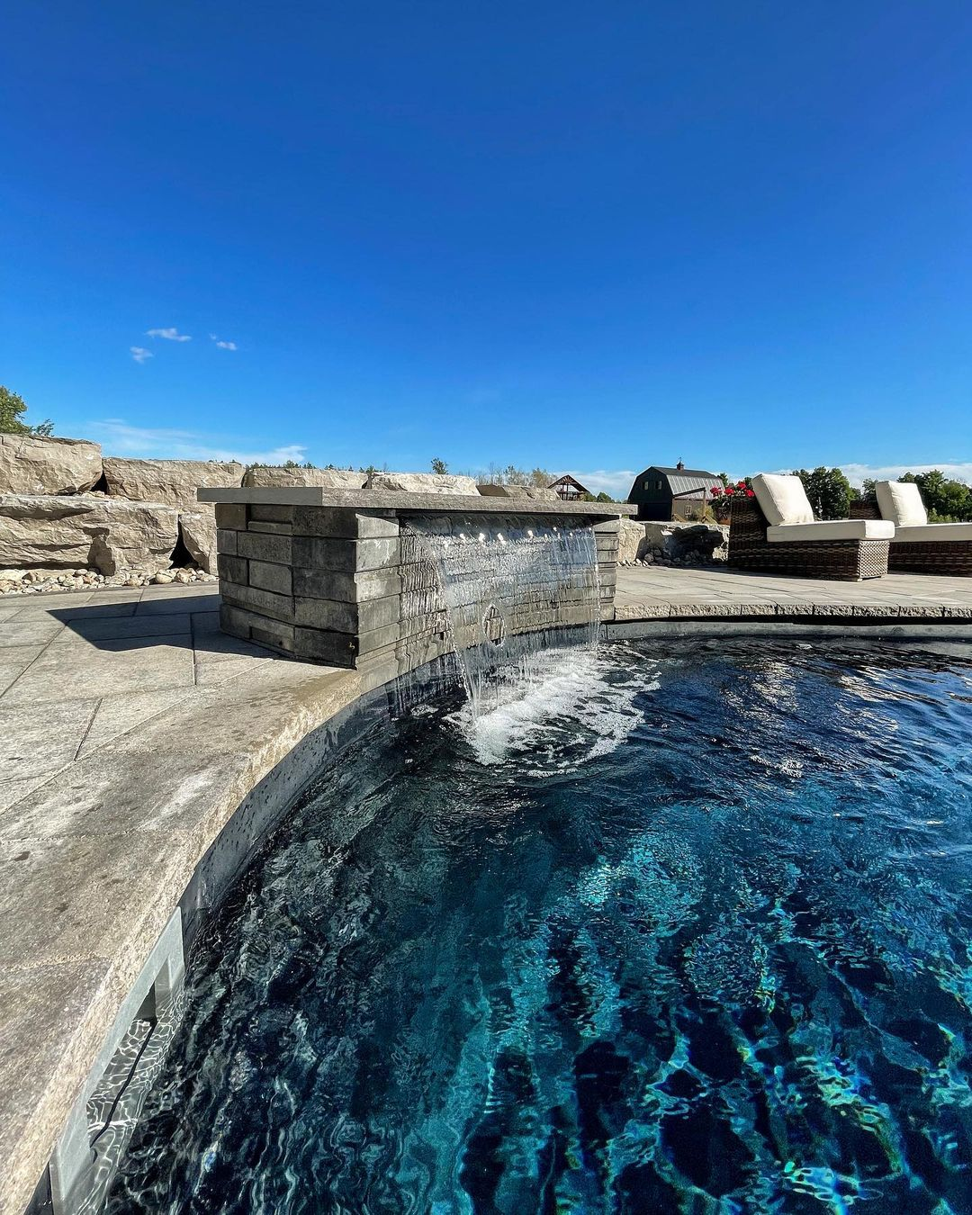 Outdoor backyard pool with a stone water feature waterfall, surrounded by lounge chairs and a clear blue sky.