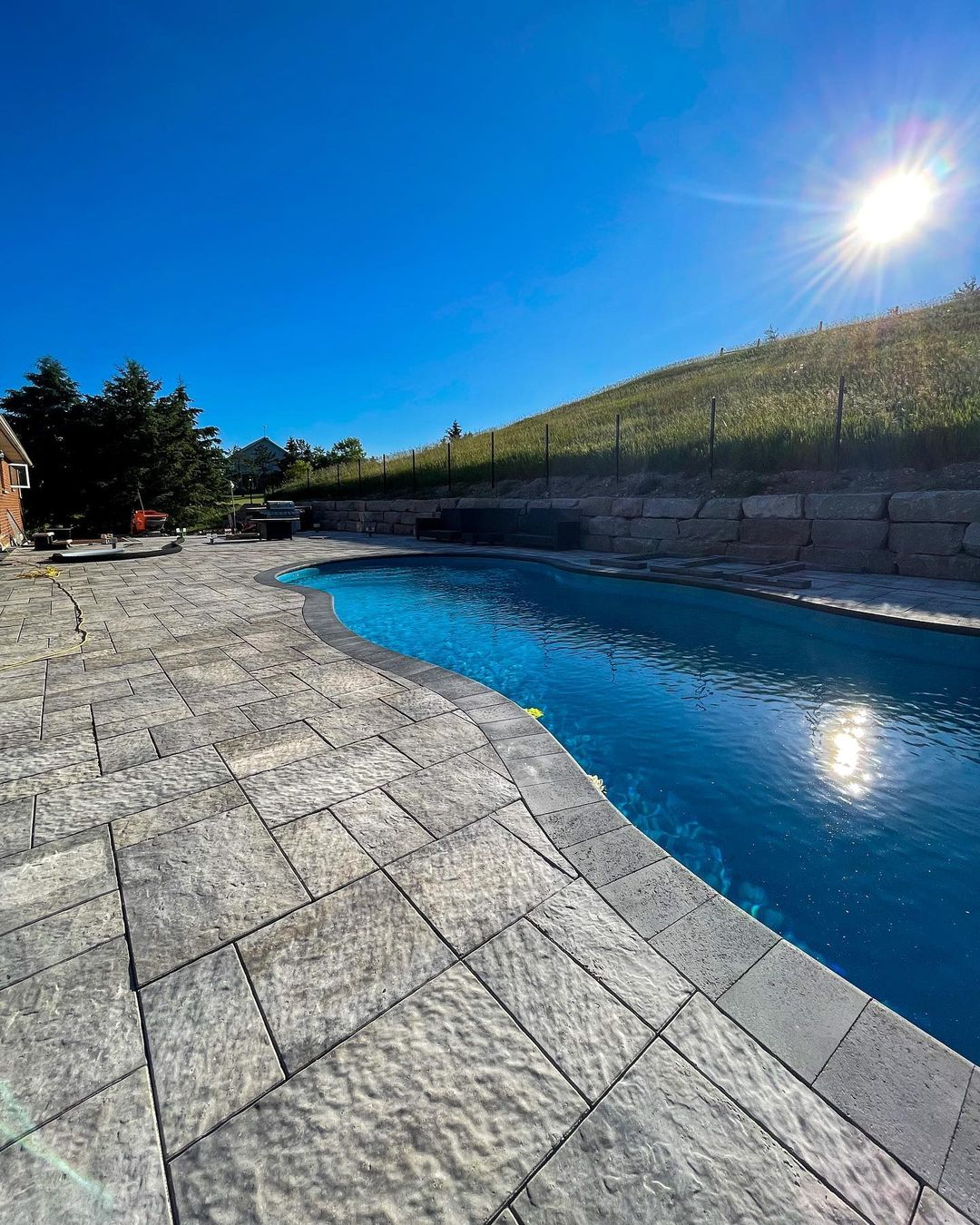 A backyard with a swimming pool surrounded by stone paving, a grassy hill in the background, and a clear blue sky with the sun shining brightly.