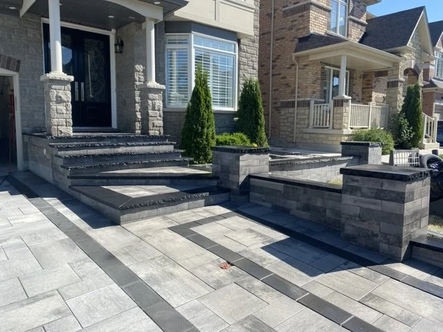 Front view of a modern house with a paved driveway, stone steps, brick and stone exterior, and tall bushes near the entrance.