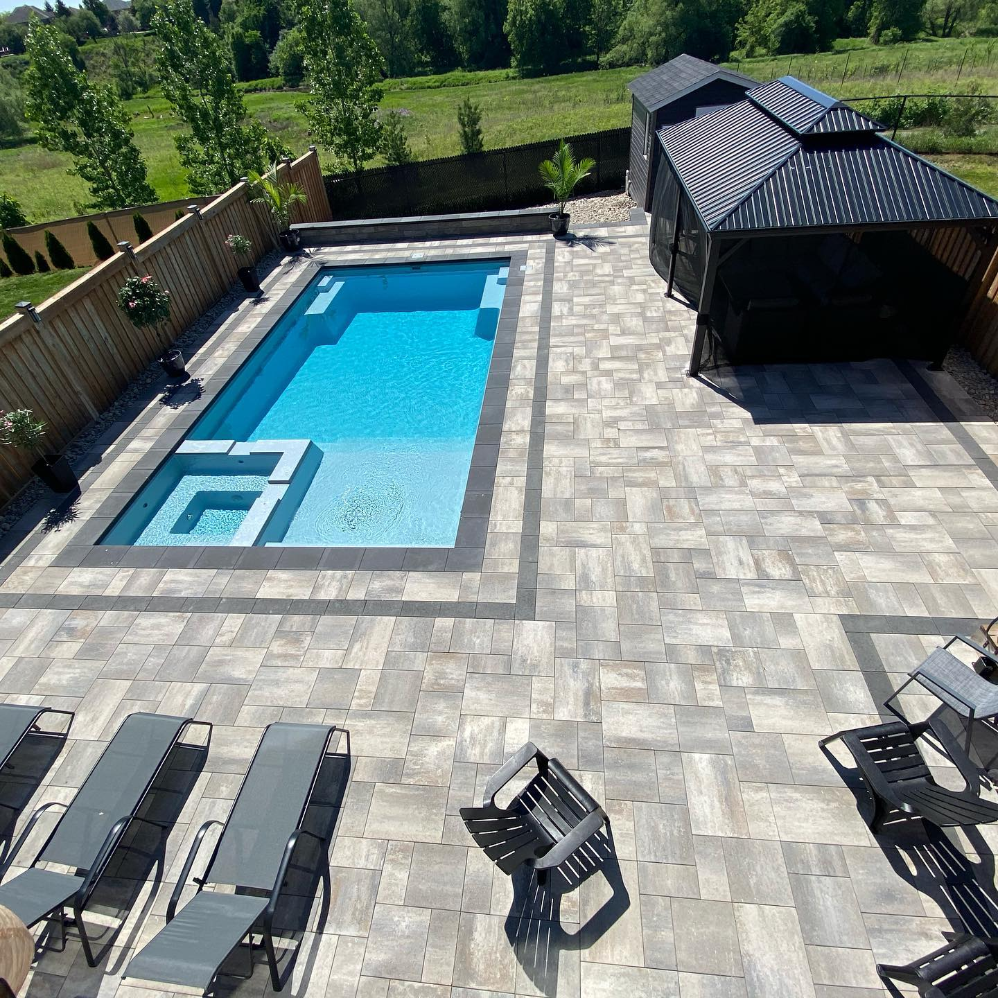 A backyard with a rectangular swimming pool, a hot tub at one end, wedding, black metal lounge chairs and a covered patio with black mesh screens, surrounded by a wooden fence with trees in the background.