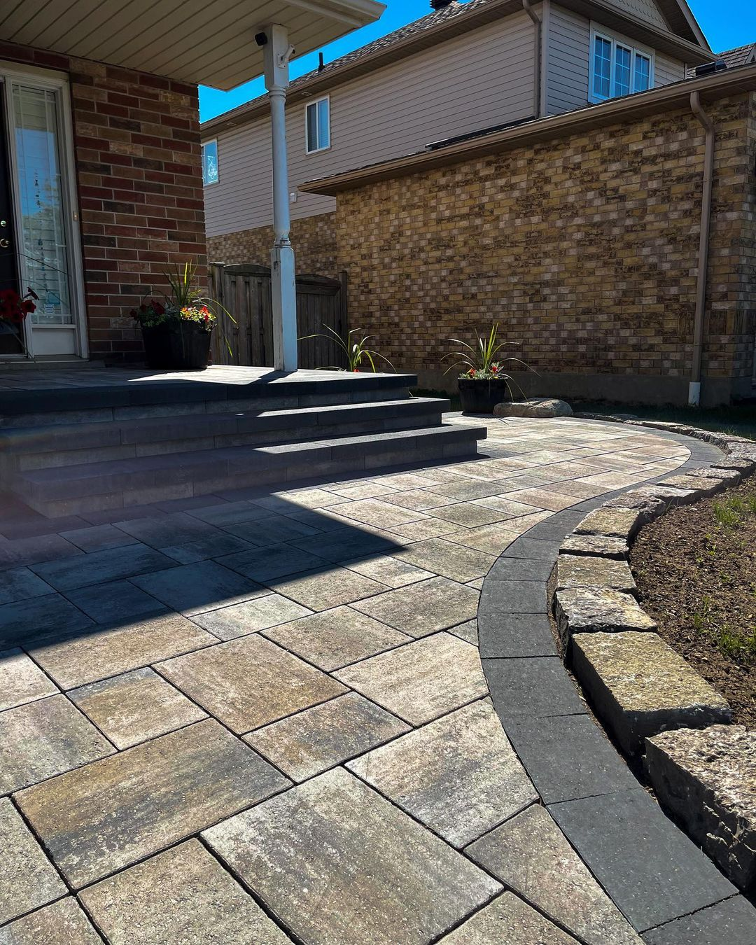 A backyard patio with stone pavers, a curved edging with larger stones, flower pots with plants, and steps leading up to a house door under a covered porch. The house has brick and siding exterior walls.