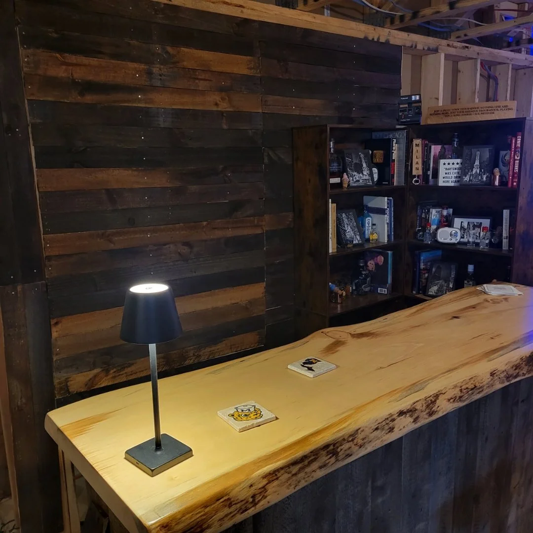 A wooden table with a black desk lamp and playing cards on it, set against a dark wooden wall and bookshelf filled with books and decorative items.
