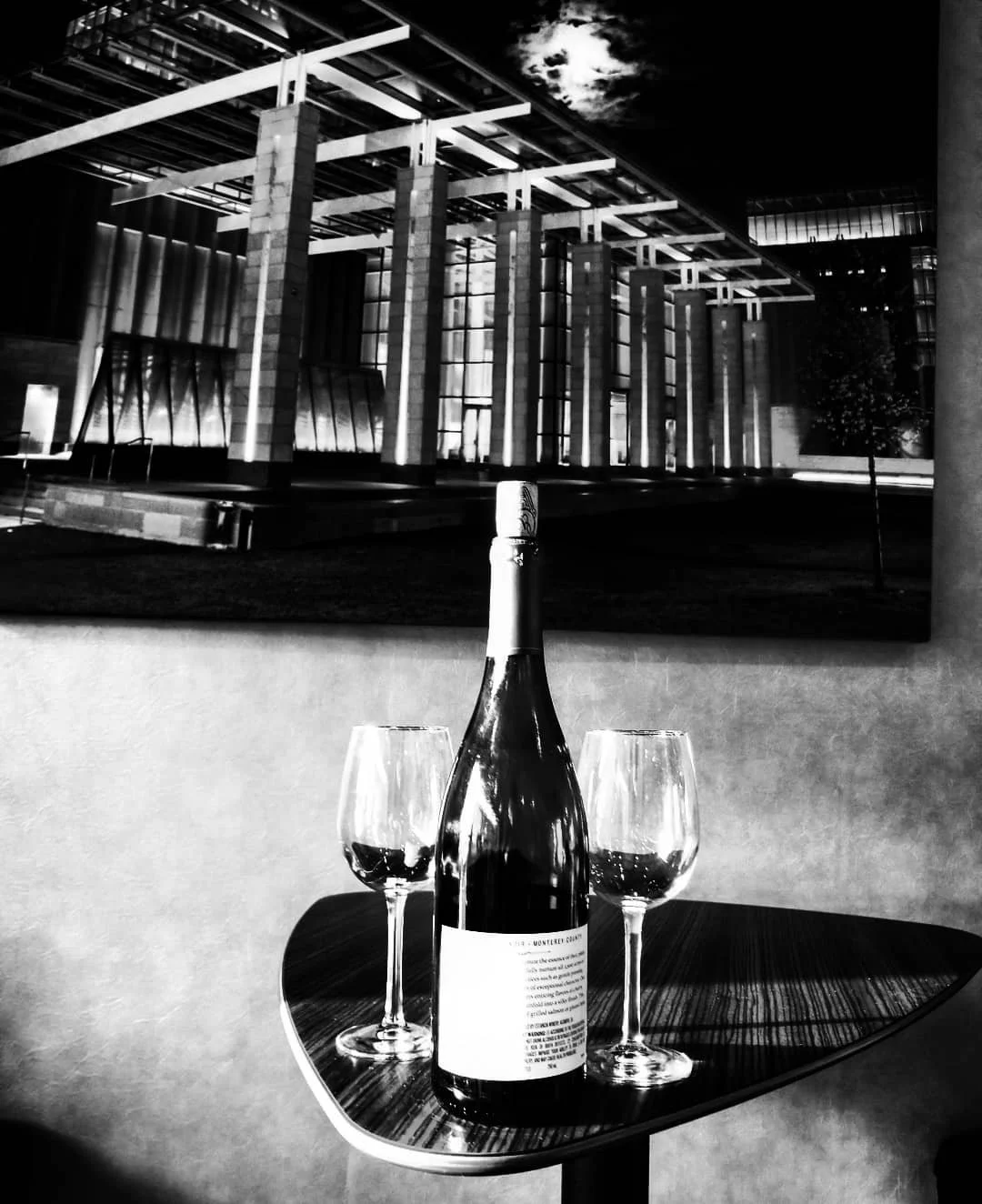 A bottle of wine and two wine glasses on a small table in front of a black-and-white photograph of a building with modern architecture at night.