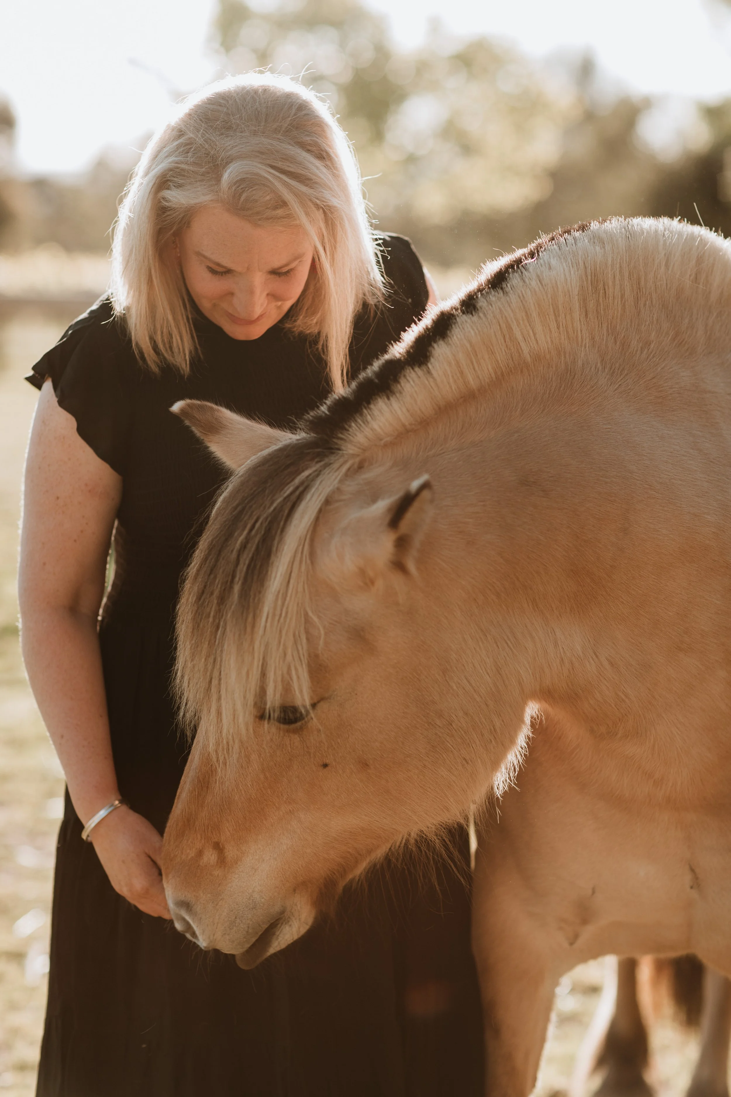 A woman with blond hair in a black dress is gently touching and looking at a light brown Fjord horse with a dark mane, outdoors with sunlight and trees in the background.