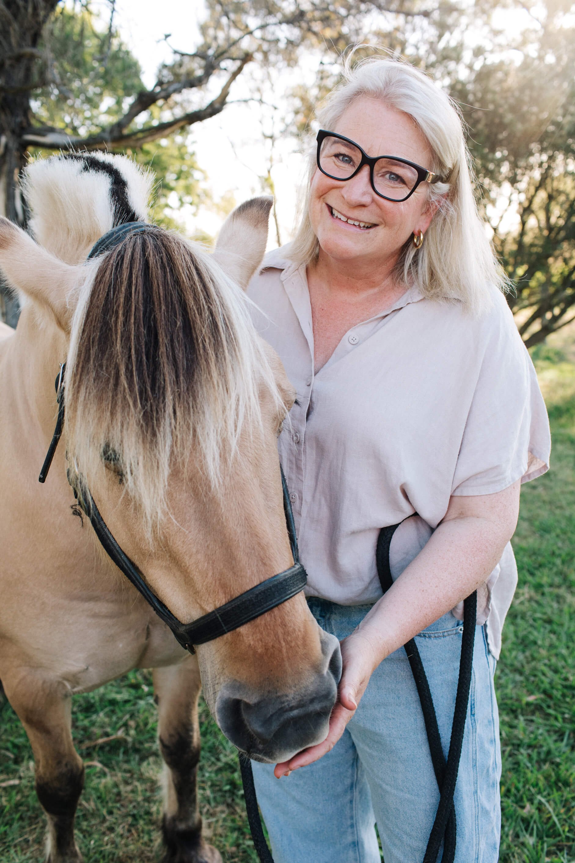 A smiling woman with blonde hair, wearing glasses, a light-colored shirt, and jeans, standing outdoors with a horse.