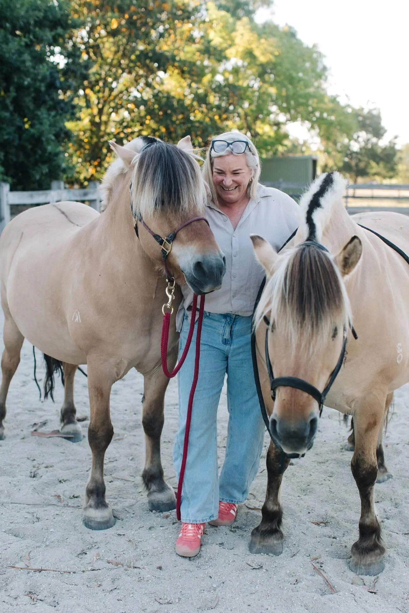 A woman smiling and standing between two Fjord horses, holding their reins, in an outdoor setting with trees and a fence in the background.