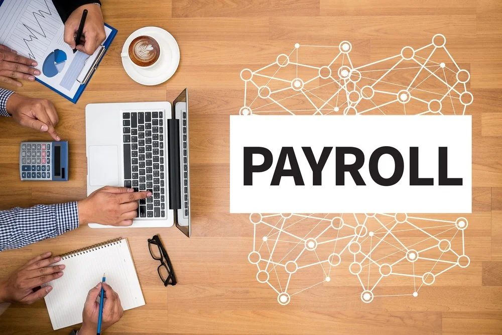 Top-down view of a workspace with a laptop, calculator, notepad, glasses, and coffee, with the word PAYROLL in bold on the right side, over a network graphic background.
