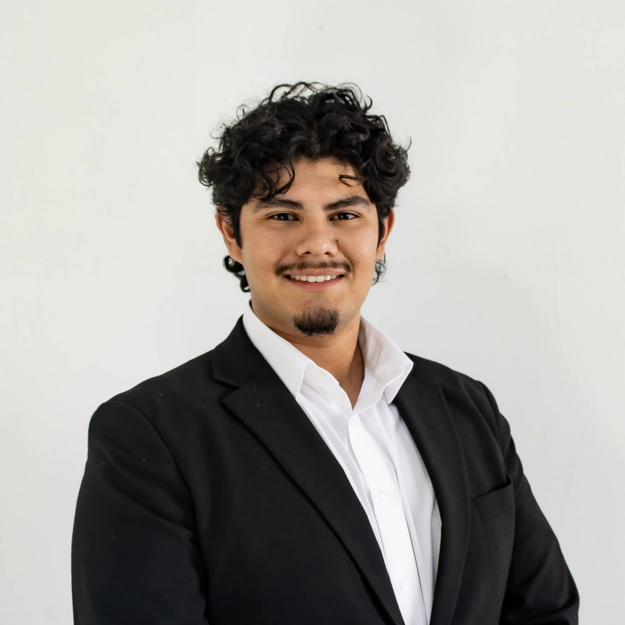 Close-up of a man with dark curly hair, smiling, wearing a black suit and white shirt, against a plain white background.