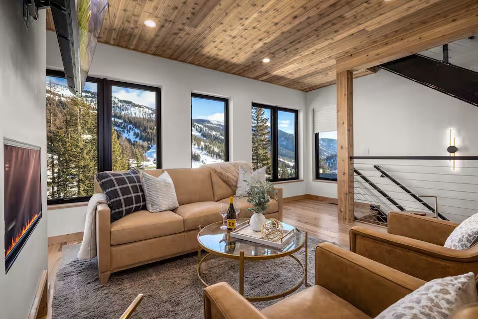 Living room with large windows showing snowy mountain landscape, beige sofa with throw pillows, glass coffee table with decorative items, wooden ceiling and floor, staircase railing, wall-mounted TV, wall light, and a fireplace.