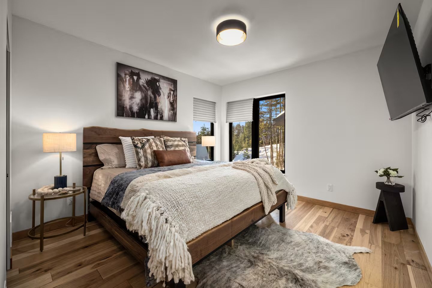 Modern bedroom with wooden bed, cozy blankets, pillows, side lamps, and large windows showing trees and snow outside.