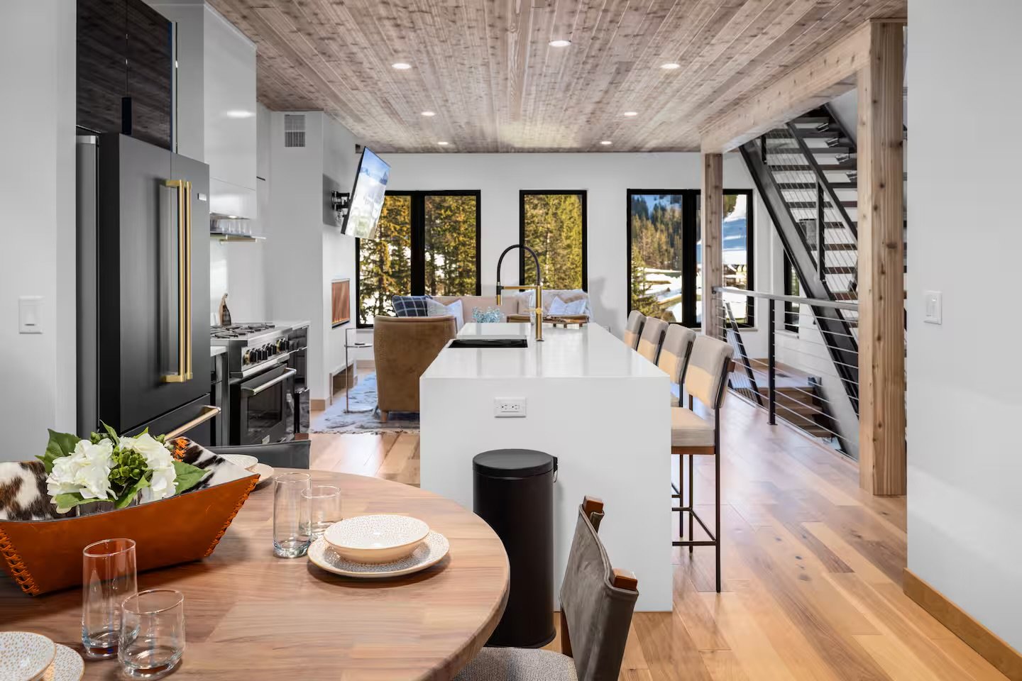 Open-concept kitchen and living area with wooden floors and ceiling, white walls, large windows with scenic views, stainless steel appliances, white kitchen island with barstools, wooden dining table with chairs, and staircase with metal railing.