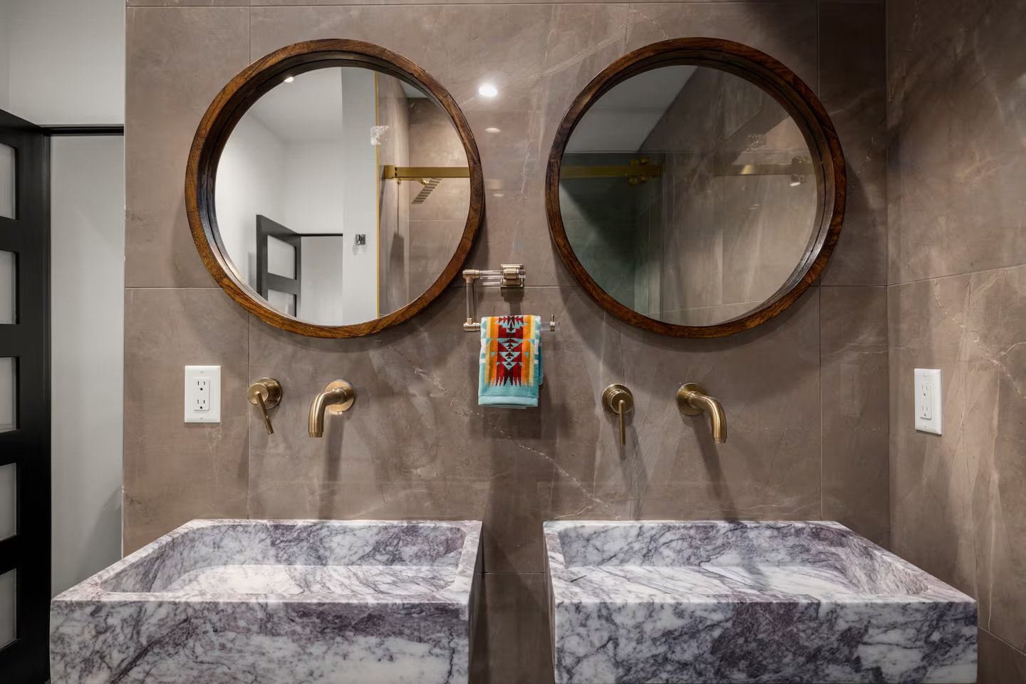 Double vanity bathroom with two oval mirrors framed in dark wood, paired with brass faucets, marble sinks, a colorful hand towel with geometric patterns, and beige tiled walls.