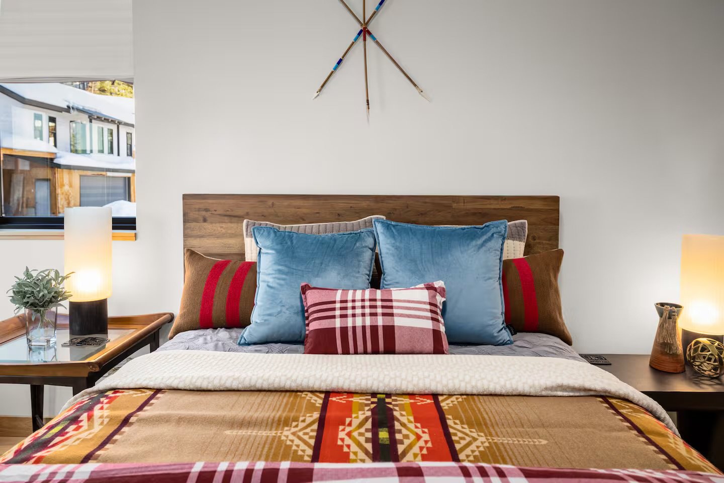 A neatly made bed with a wood headboard, decorated with blue, red, brown, and patterned pillows, positioned between two bedside tables with lamps, in a bedroom with a window and a minimalistic wall decoration.