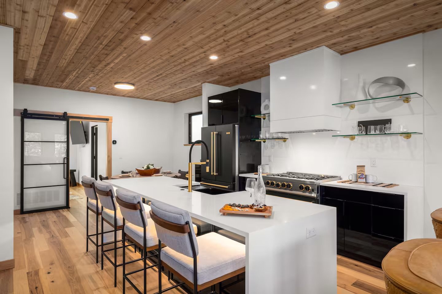 Modern kitchen with white island, black cabinetry, wooden ceiling, open shelving, and seating area with six chairs.